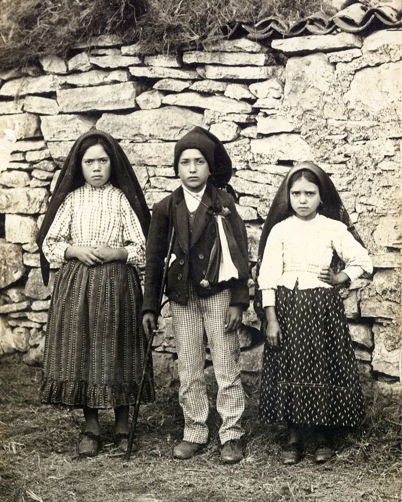 Fotografia antiga em tons de sépia de três crianças em pé diante de um muro de pedra: duas meninas de véu escuro e um menino de chapéu ao centro.