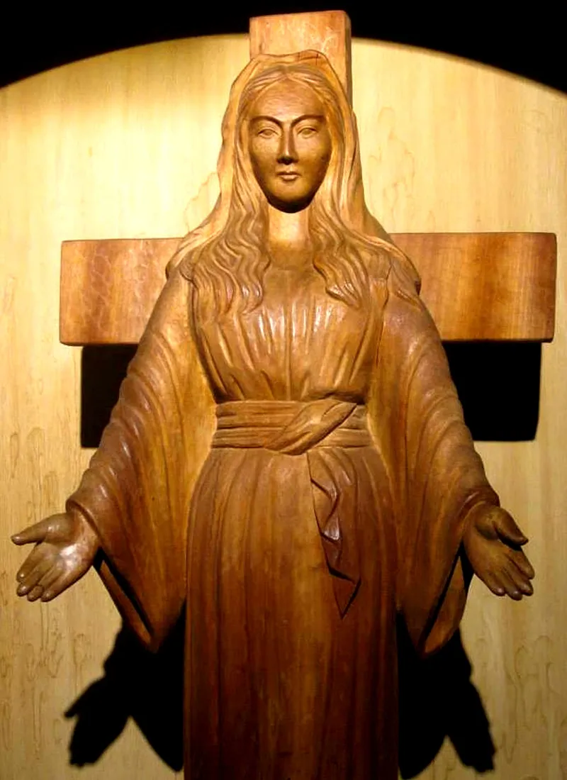Escultura de madeira de Maria diante de uma cruz, com os braços abertos para baixo.