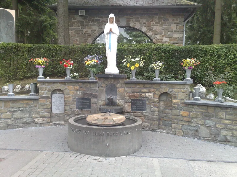 Pequena estátua branca de Maria num nicho de pedra, acima de uma fonte, com flores ao redor.