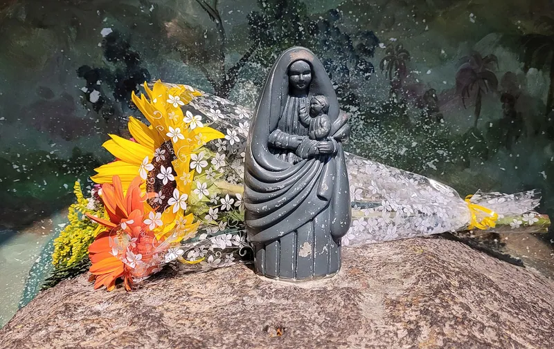 Pequena imagem escura de Maria com o Menino, sobre uma pedra, com flores ao lado.