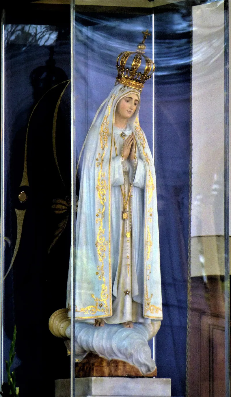 Estátua de Maria vestida de branco com bordas douradas, mãos postas e terço pendente, sob uma coroa.
