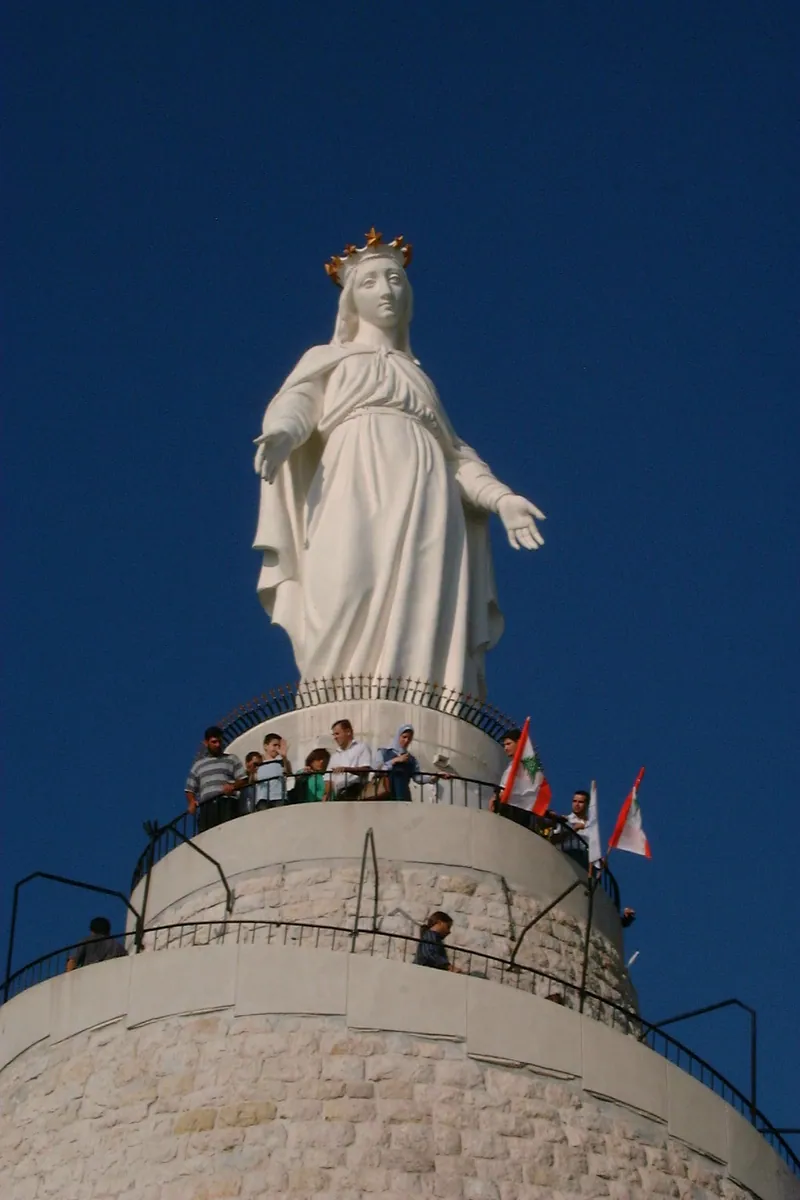 Grande estátua branca de Maria de braços abertos sobre um pedestal, contra o céu.