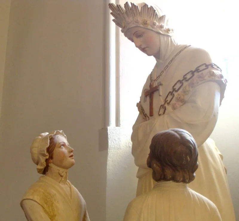 Estátua de Maria com coroa de rosas, crucifixo e corrente sobre o peito, diante de duas crianças.