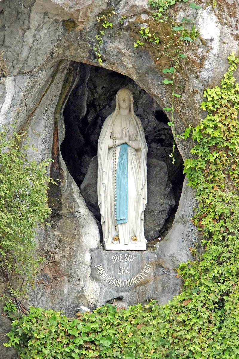 Estátua branca de Maria com faixa azul, mãos postas, no alto de um nicho na rocha da gruta.