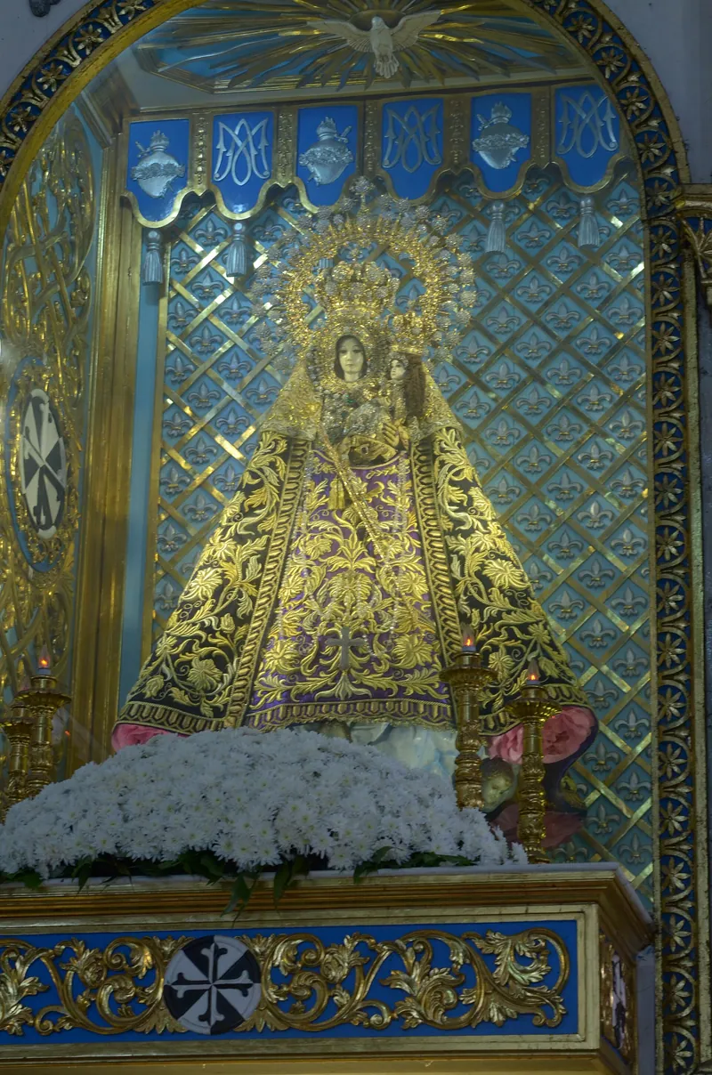 Imagem de Maria com vestes douradas e coroa, num nicho ricamente ornamentado com flores brancas à frente.