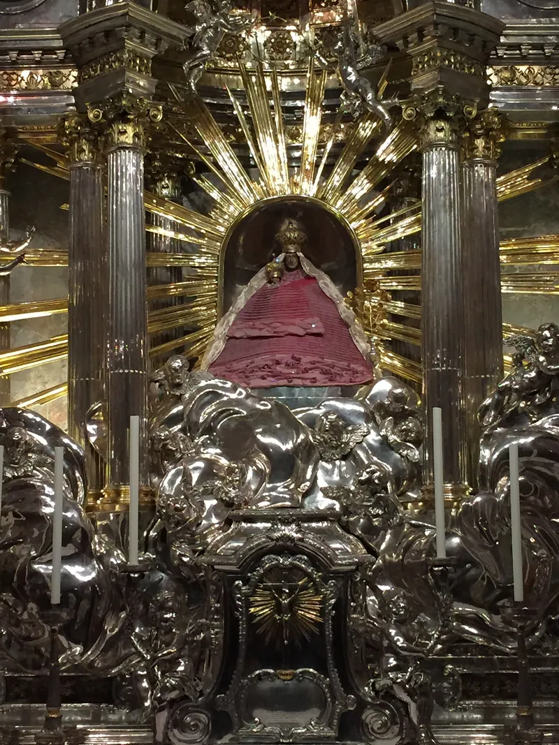 Pequena imagem de Maria com manto vermelho, num altar de prata cercado de colunas douradas.